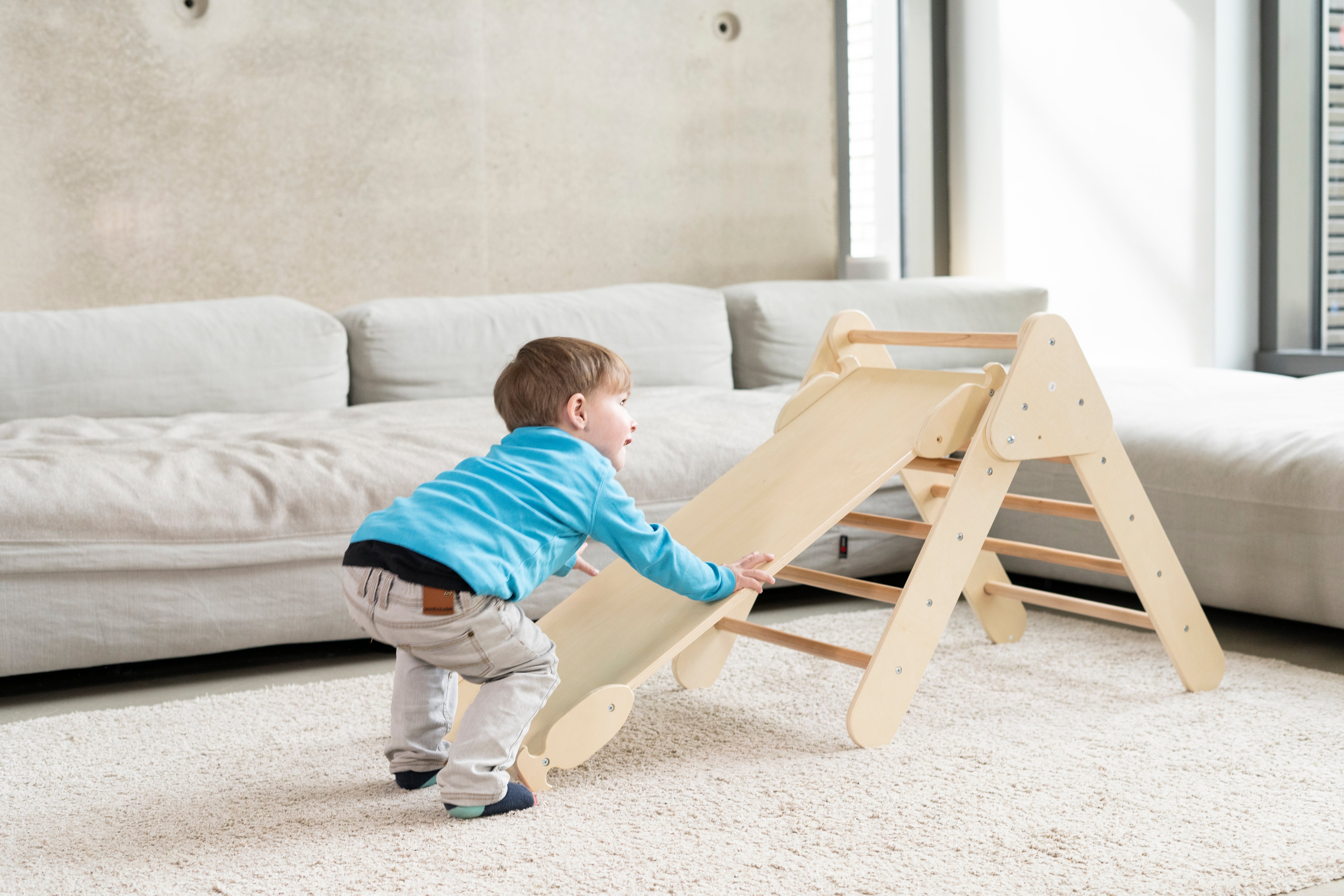 Kleiner Junge spielt an einem stabilen Kletterdreieck mit Rutsche aus Holz im Wohnzimmer. Das Kletterdreieck fördert motorische Fähigkeiten und bietet sicheren Spielspaß für Kinder.