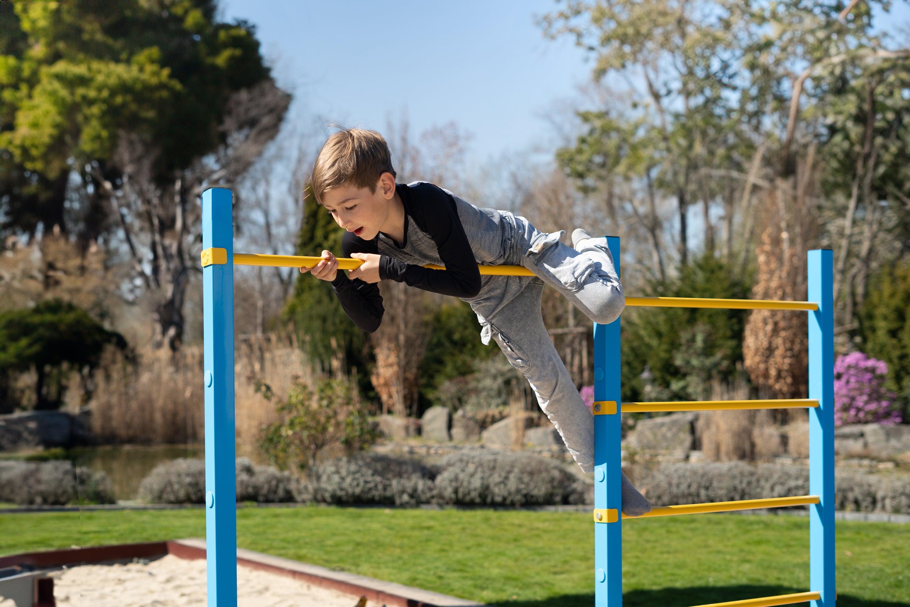 Kind balanciert spielerisch auf einer höhenverstellbaren Reckstange eines bunten Outdoor-Turngeräts im Garten. Das stabile Klettergerüst ist ideal als Spielgerät für Kinder und fördert Bewegung im Freien.
