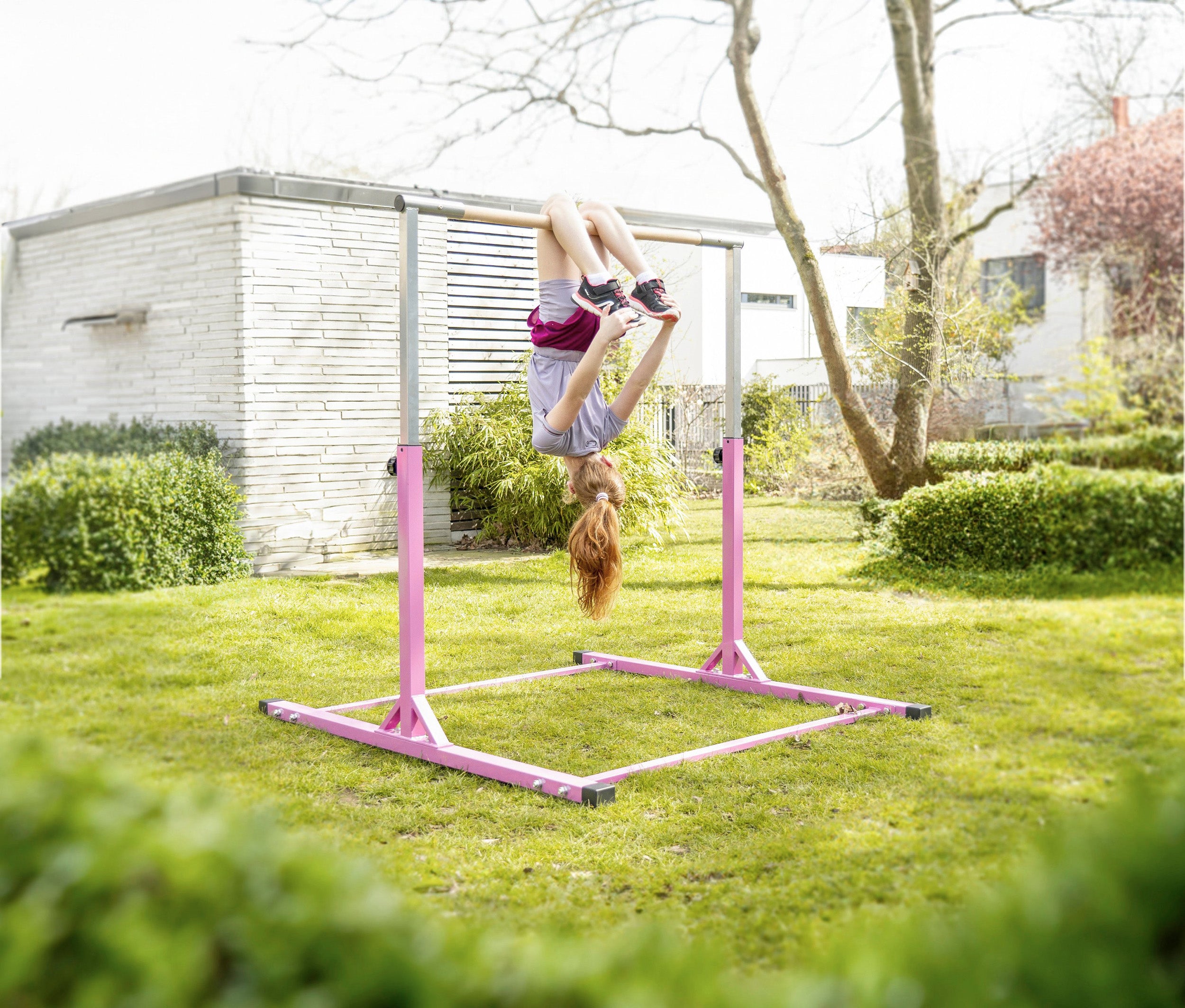 Kinderturnreck aus Metall in Pink, im Garten aufgebaut. Ein Mädchen turnt kopfüber an der Stange und zeigt Spaß an der Bewegung. Das Turnreck steht stabil auf einer grünen Rasenfläche.