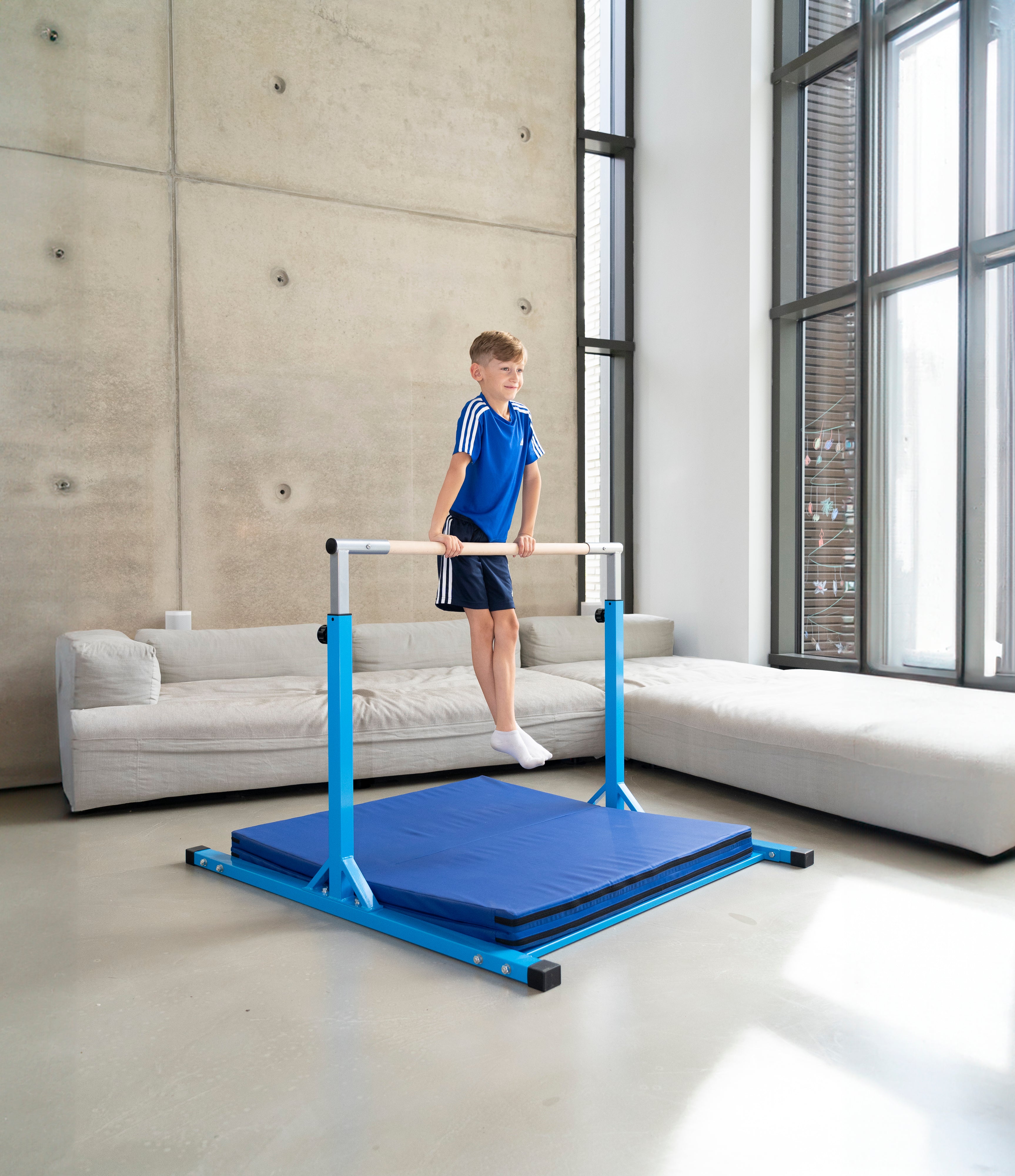 Junge trainiert an einem höhenverstellbaren Indoor-Turnreck mit weicher Bodenmatte in einem modernen Wohnzimmer. Das kompakte Turngerät mit Holzstange eignet sich perfekt für Kinder zum sicheren Turnen zu Hause.
