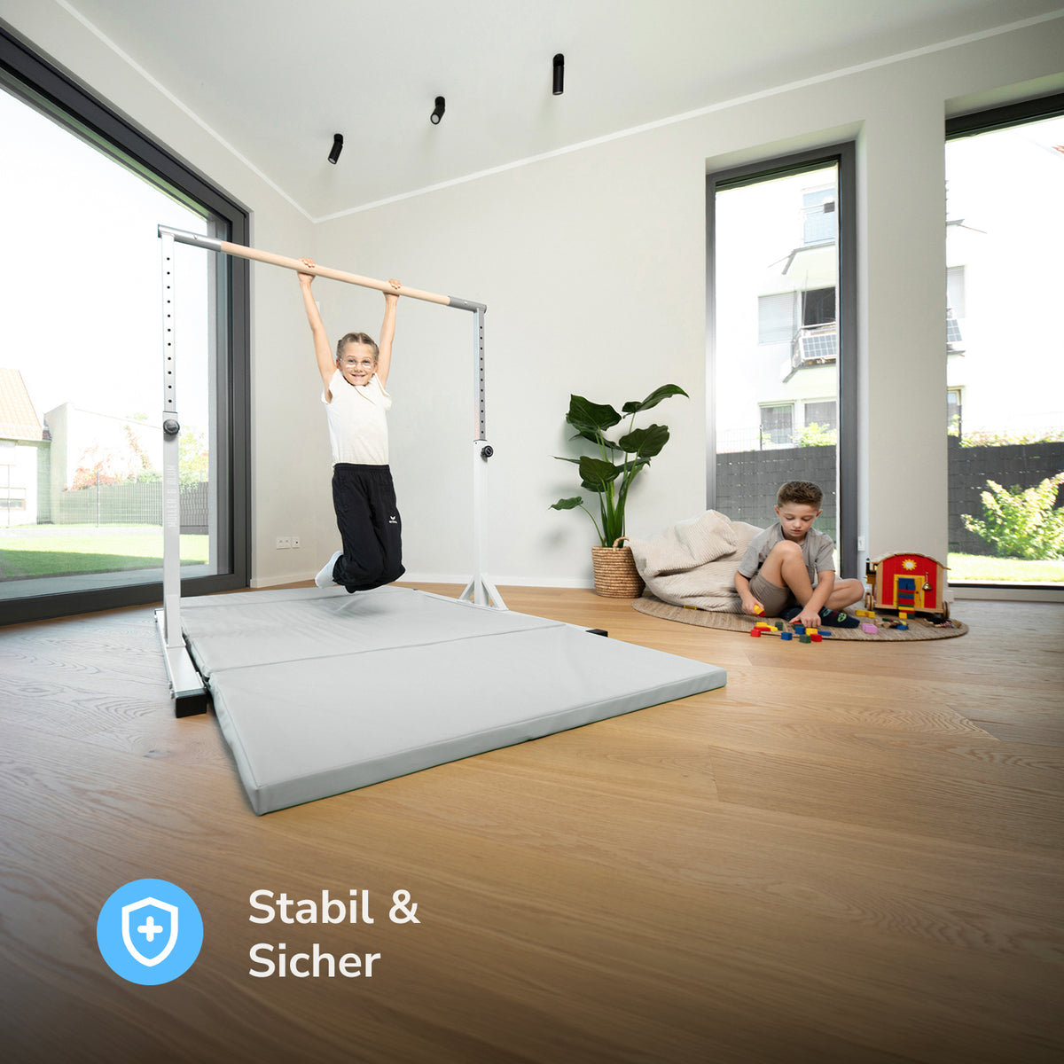 Mädchen hängt an einem höhenverstellbaren Turnreck in Warmgrau mit Turnmatte im Wohnzimmer, daneben spielt ein Junge mit Bauklötzen