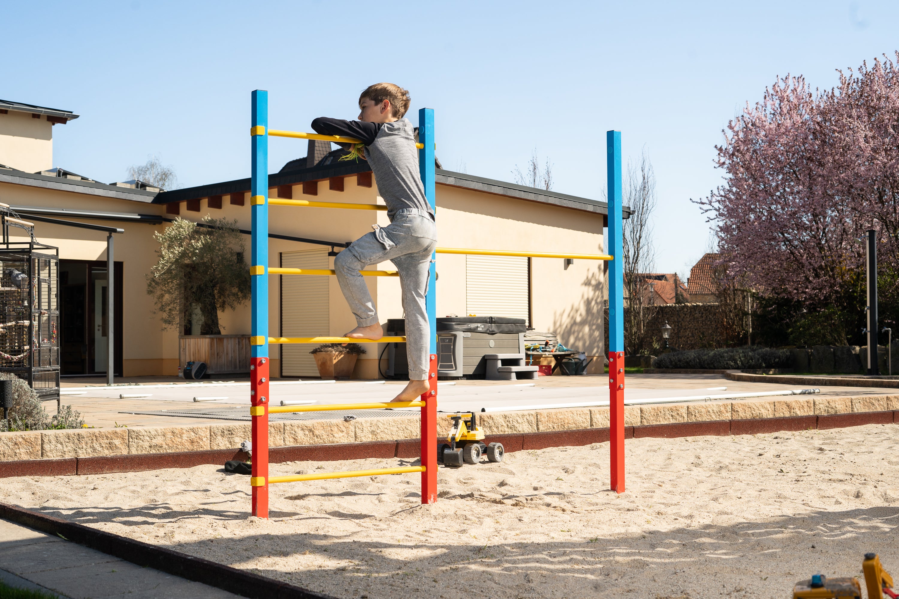 Barfuß klettert ein Junge an einem farbenfrohen Outdoor-Turnreck mit Sprossenwand und Reckstange im Garten. Das Spielgerät aus Stahl steht sicher im Sandbereich und eignet sich ideal zum Klettern und Turnen für Kinder.