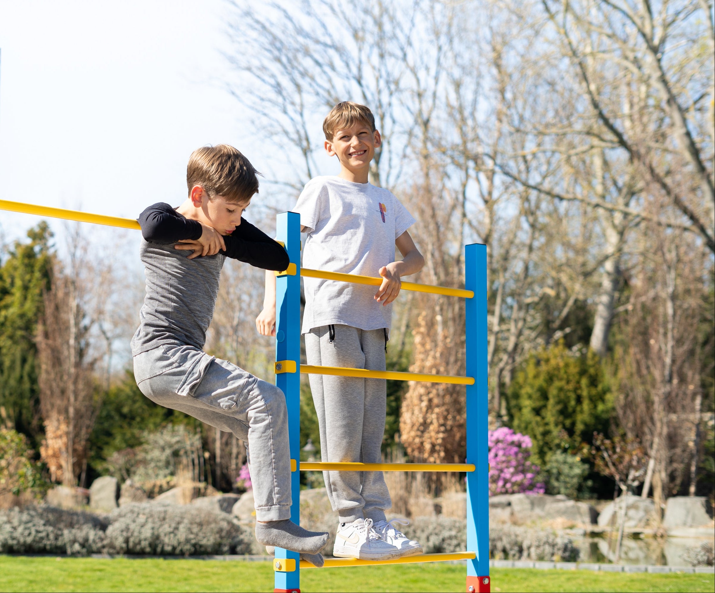 Zwei Jungen spielen auf einem farbenfrohen Outdoor-Klettergerüst im Garten. Einer balanciert an der höhenverstellbaren Reckstange, während der andere auf der Sprossenwand steht. Das robuste Turnreck bietet vielfältige Spiel- und Bewegungsmöglichkeiten für Kinder.