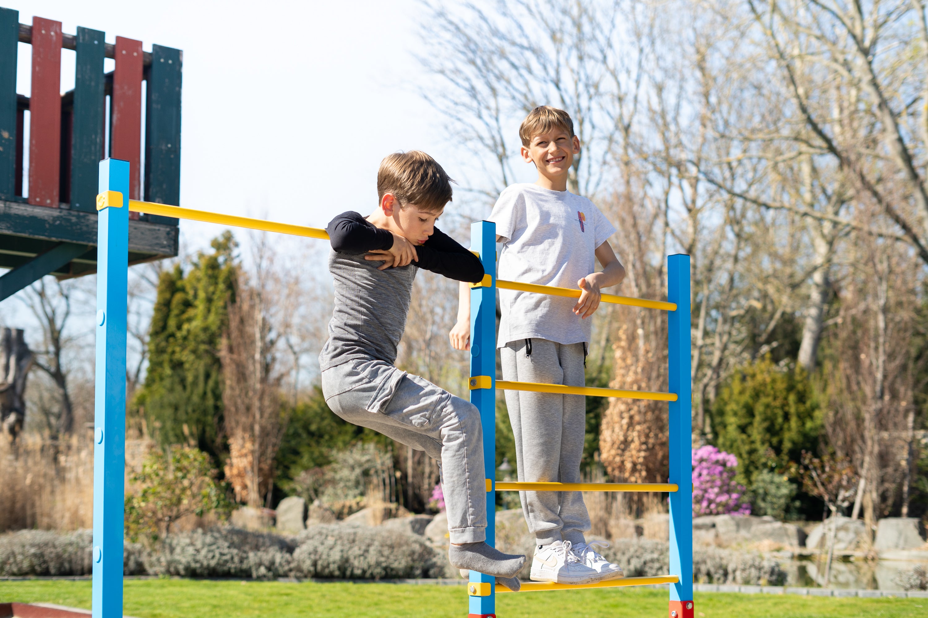 Zwei Jungen spielen auf einem farbenfrohen Outdoor-Klettergerüst im Garten. Einer balanciert an der höhenverstellbaren Reckstange, während der andere auf der Sprossenwand steht. Das robuste Turnreck bietet vielfältige Spiel- und Bewegungsmöglichkeiten für Kinder.